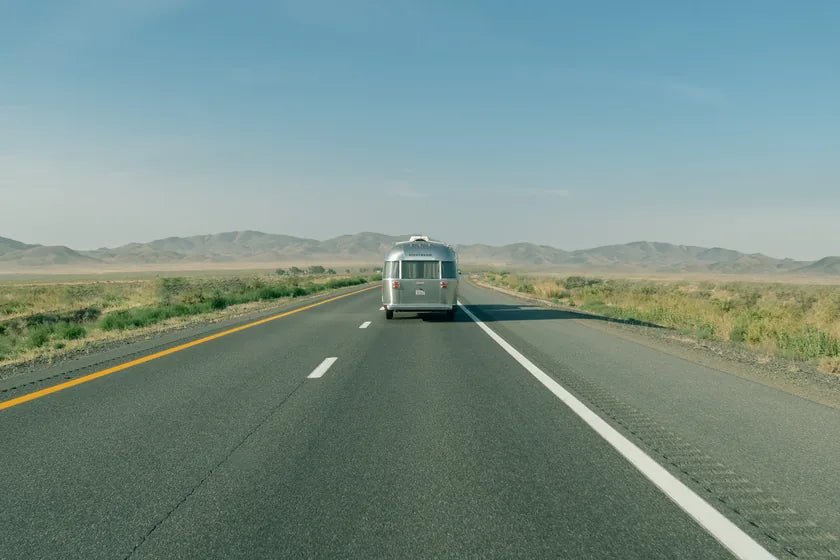 A silver travel trailer on an open highway toward distant mountains
