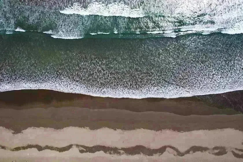 An aerial view of pale sand and foam meeting open water — the lived-in coast as pattern, not place.