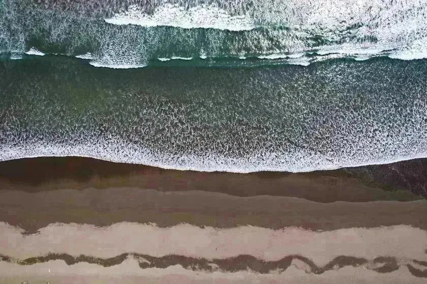 An aerial view of pale sand and foam meeting open water — the lived-in coast as pattern, not place.