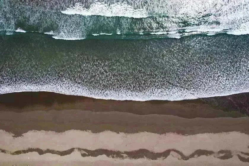 An aerial view of pale sand and foam meeting open water — the lived-in coast as pattern, not place.